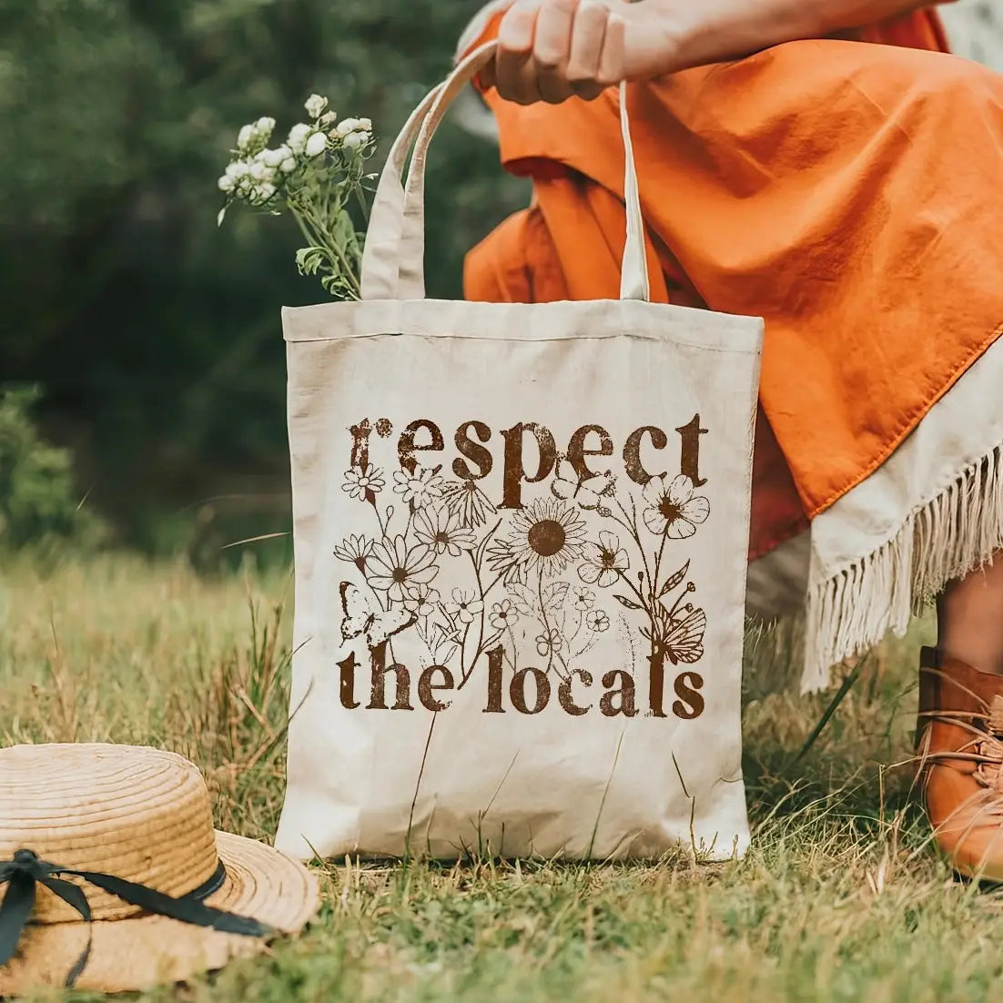 Person holding a tote bag with 'respect the locals' text in a grassy outdoor setting