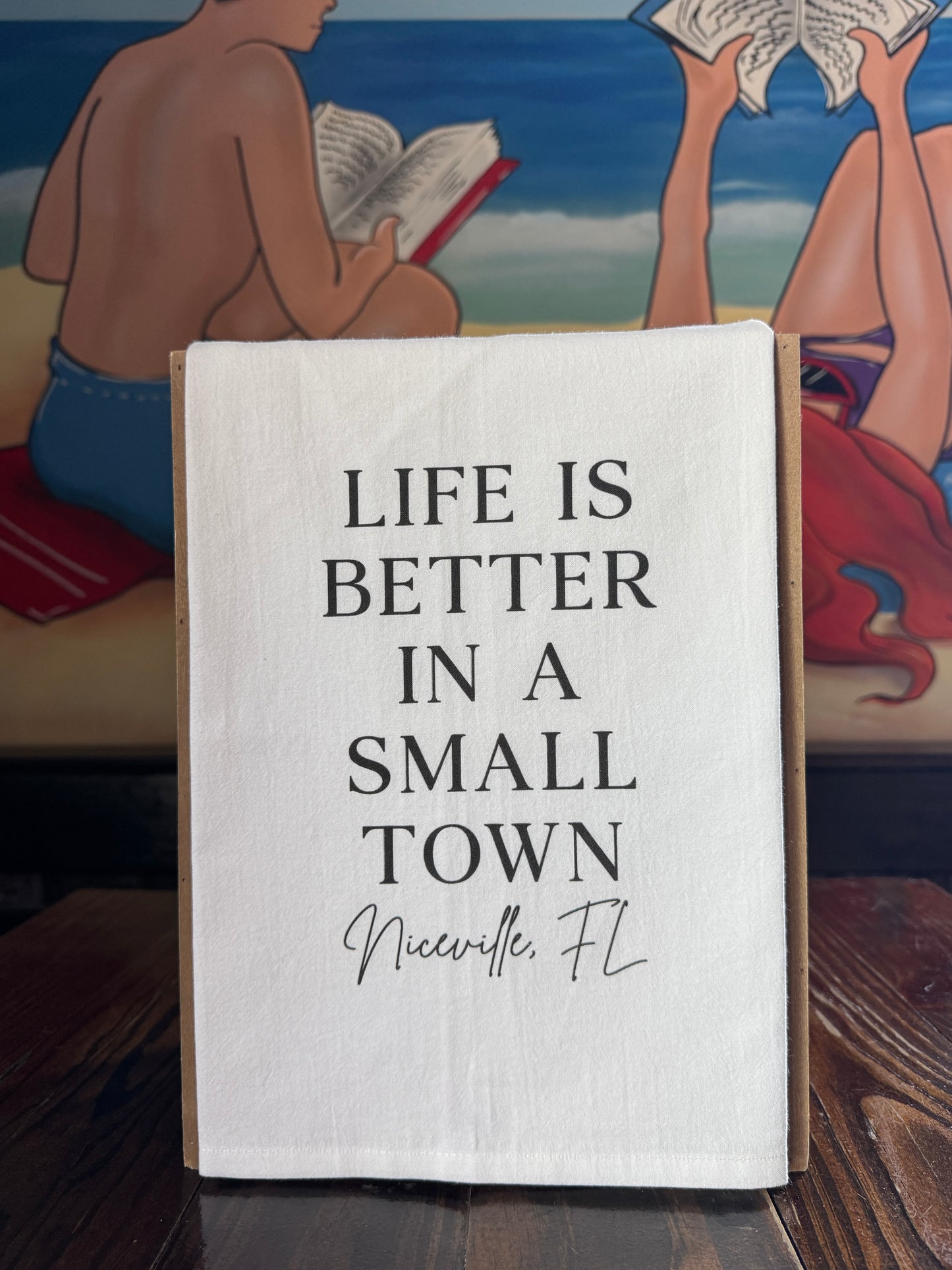 Textile with 'Life is better in a small town Niceville, FL' on a wooden surface with a mural in the background.