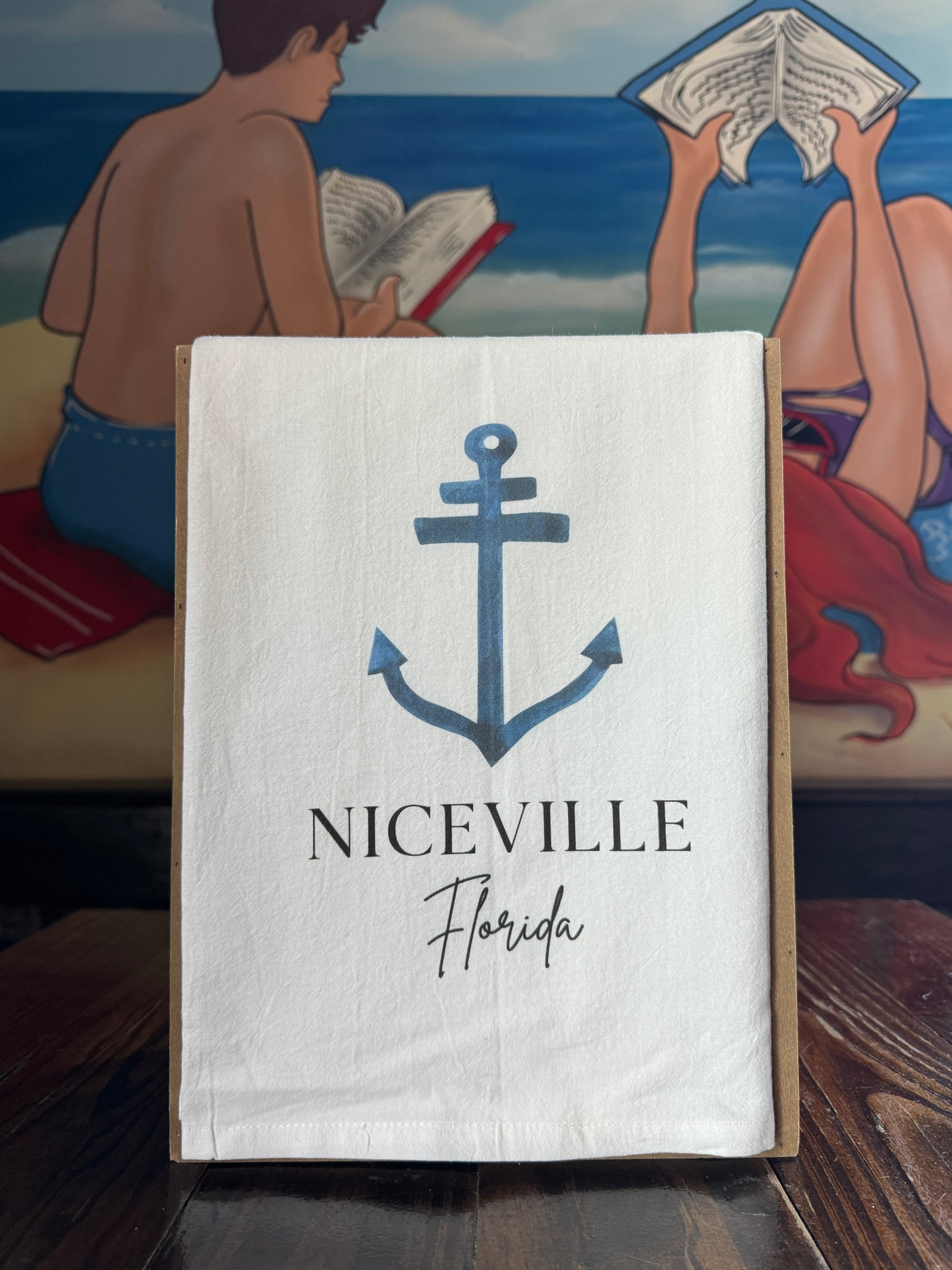 White towel with anchor design and 'Niceville Florida' text, placed on a surface with a painting in the background.