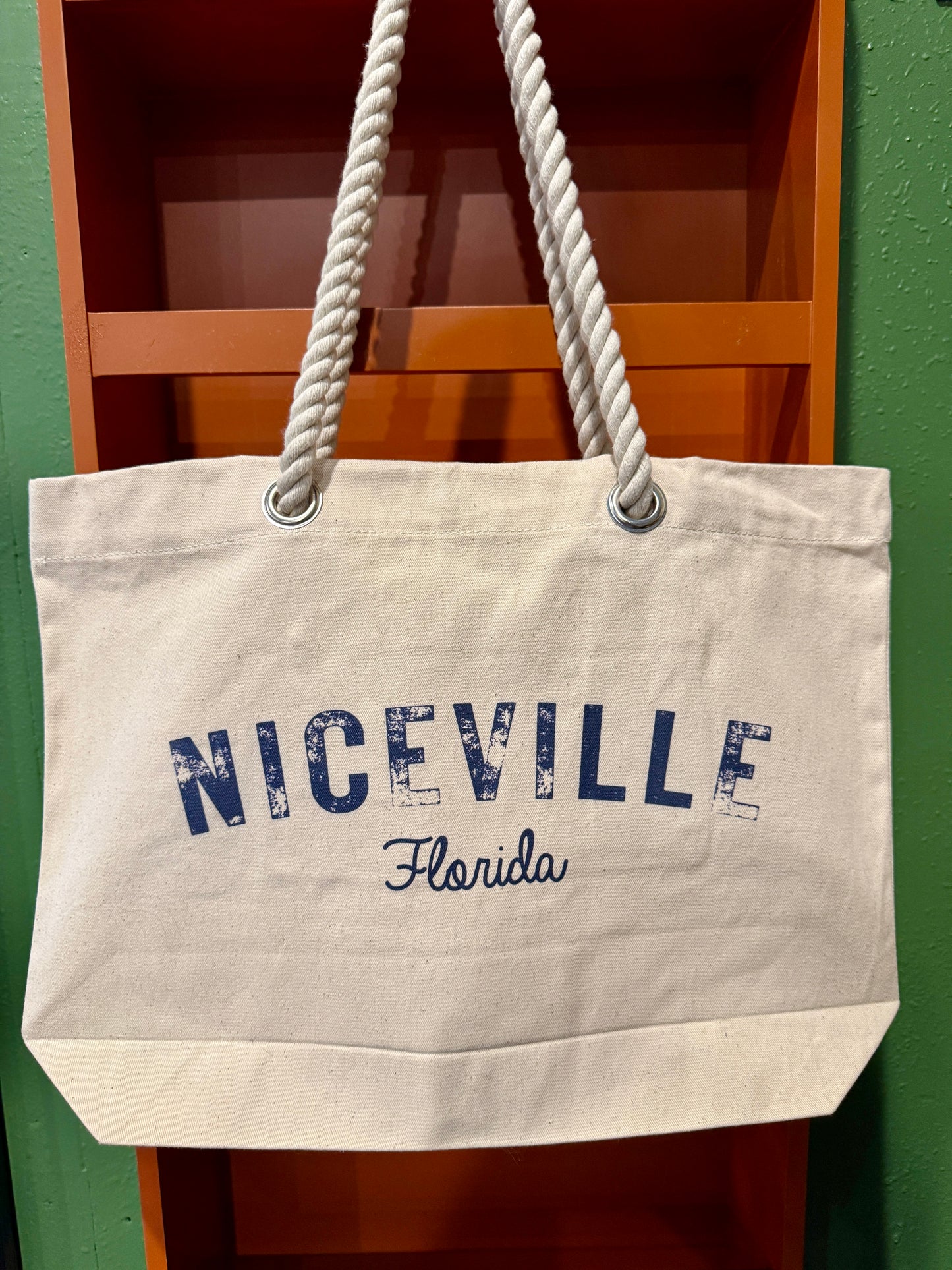 Beige tote bag with 'Niceville Florida' text on a wooden shelf against a green wall.