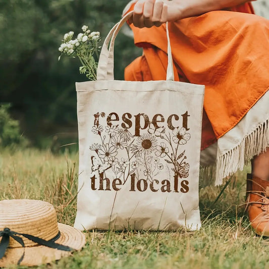 Person holding a tote bag with 'respect the locals' text in a grassy outdoor setting