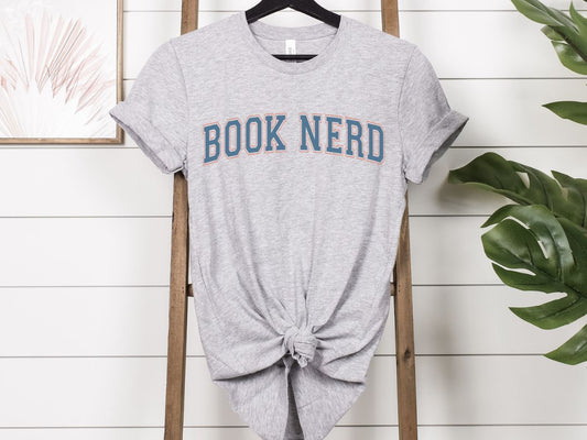 Gray t-shirt with 'BOOK NERD' text on a wooden rack against a white wall.