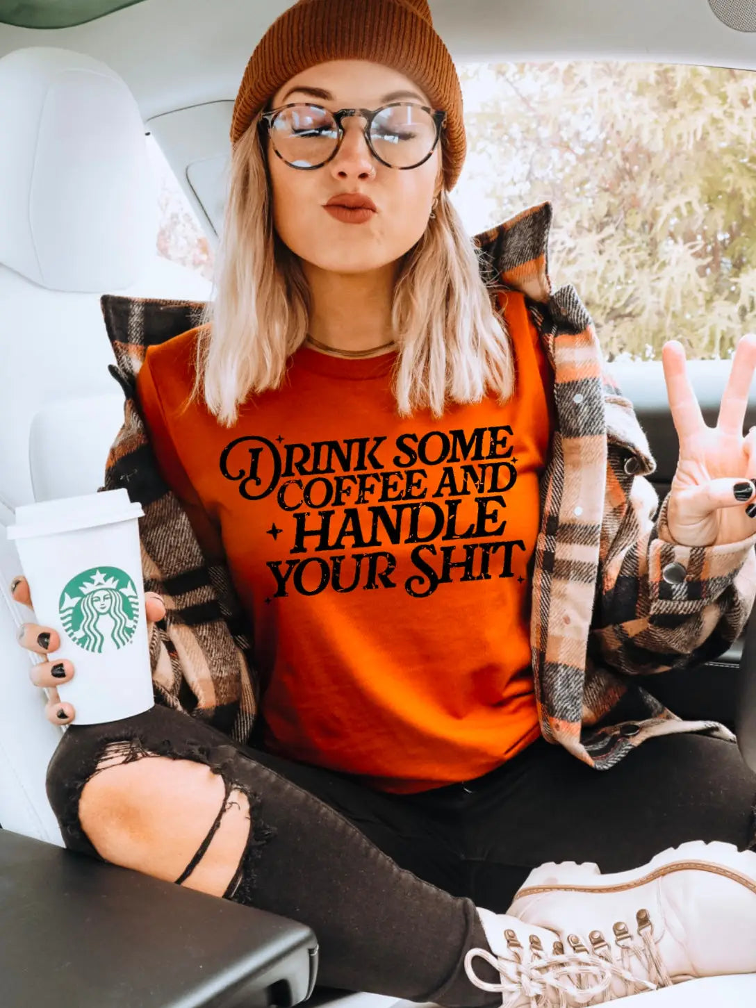 Person wearing an orange sweatshirt with text, holding a Starbucks cup, and sitting in a car.