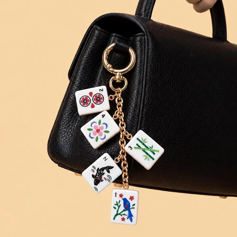 A gold-tone keychain charm featuring five white mahjong tile charms with colorful traditional Chinese symbols, hanging from a black handbag on a warm beige background.