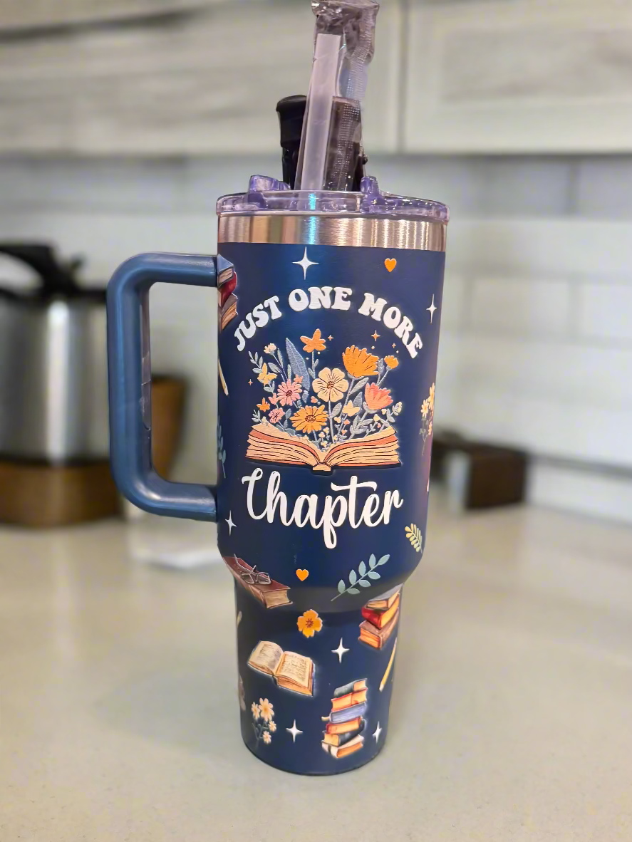 Navy blue tumbler with book-themed design and text 'Just One More Chapter' on a wooden surface.