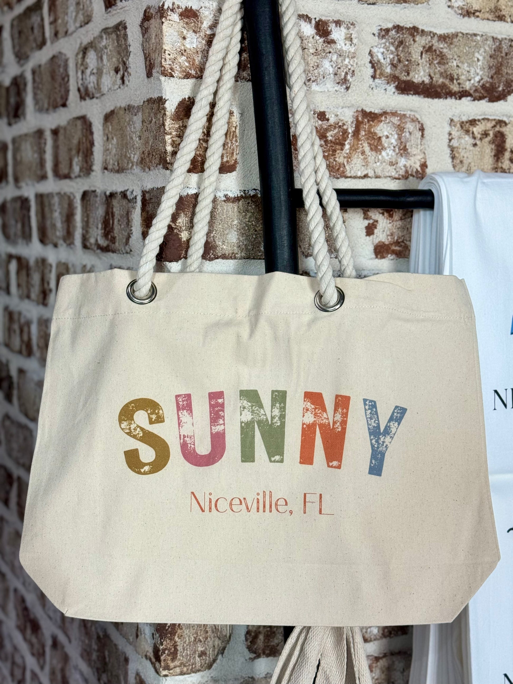 Beige tote bag with 'SUNNY Niceville, FL' text against a brick wall.