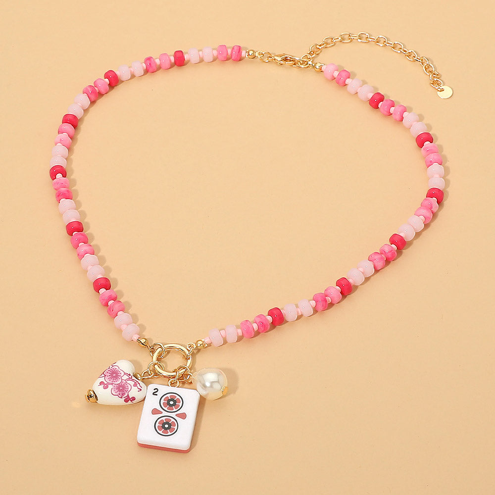 A pink and white beaded necklace with gold-tone hardware, featuring a white enamel tile charm, a small pearl bead, and a dangling floral heart charm, displayed on a solid peach background.