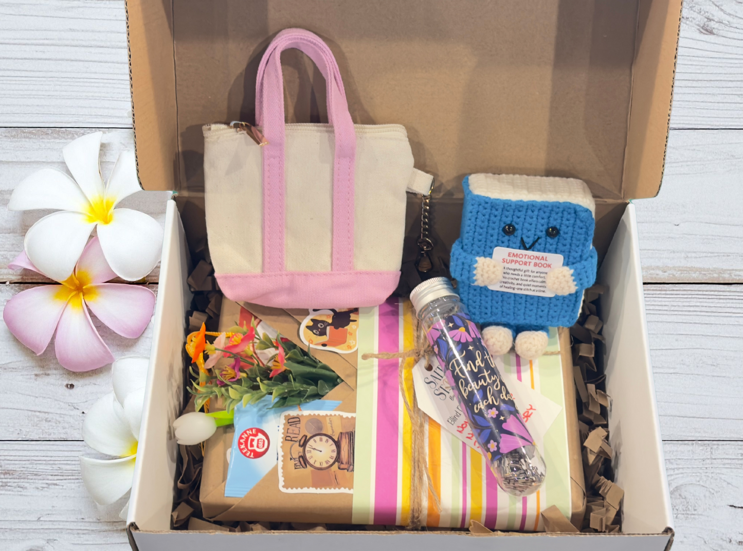 Box containing a small handbag, plush toy, and other items on a wooden surface with flowers.