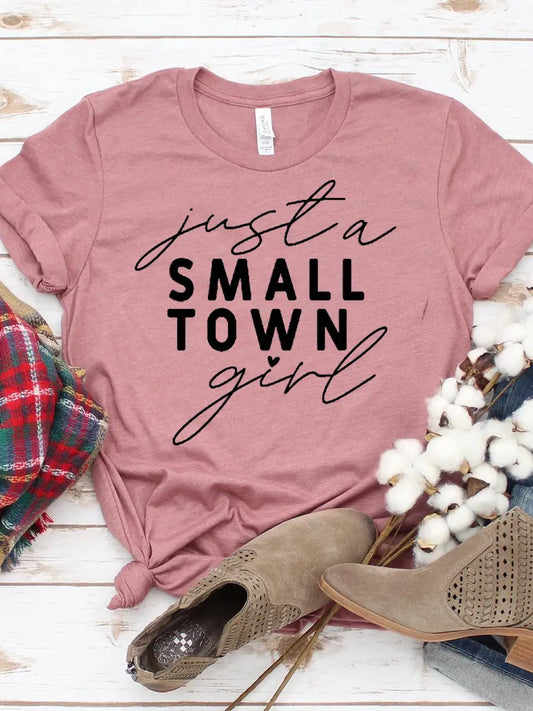 Pink t-shirt with 'just a small town girl' text, paired with boots and plaid fabric on a wooden surface.